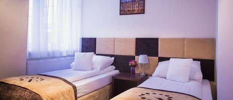 A hotel room with two beds, a nightstand, a lamp, a window with sheer curtains, and a framed picture on the wall.