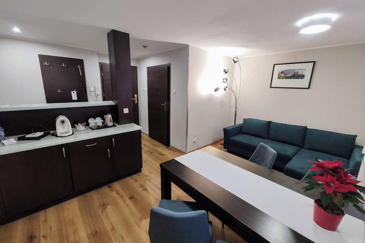 A modern kitchen with dark cabinets, a white countertop, and a wooden floor. There is a dining table with chairs and a potted plant. A teal sofa is against the wall, and a floor lamp is visible.