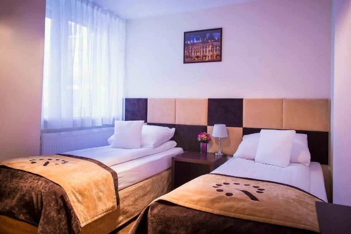 A hotel room with two beds, a nightstand, a lamp, a window with sheer curtains, and a framed picture on the wall.