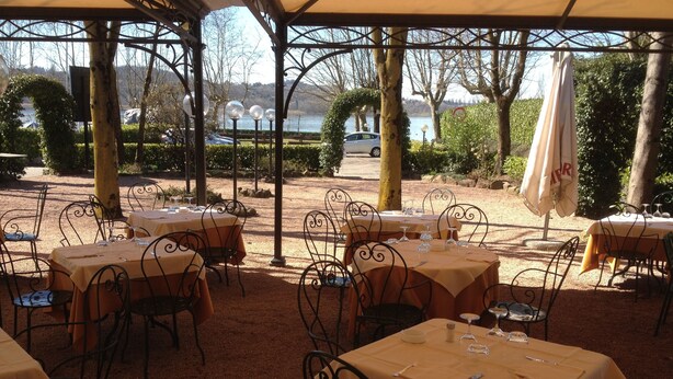 An outdoor dining area with tables and chairs, a covered section with a view of a lake and hills, and a parking area in the background. - Hotel Ristorante Vecchia Riva (Varese)