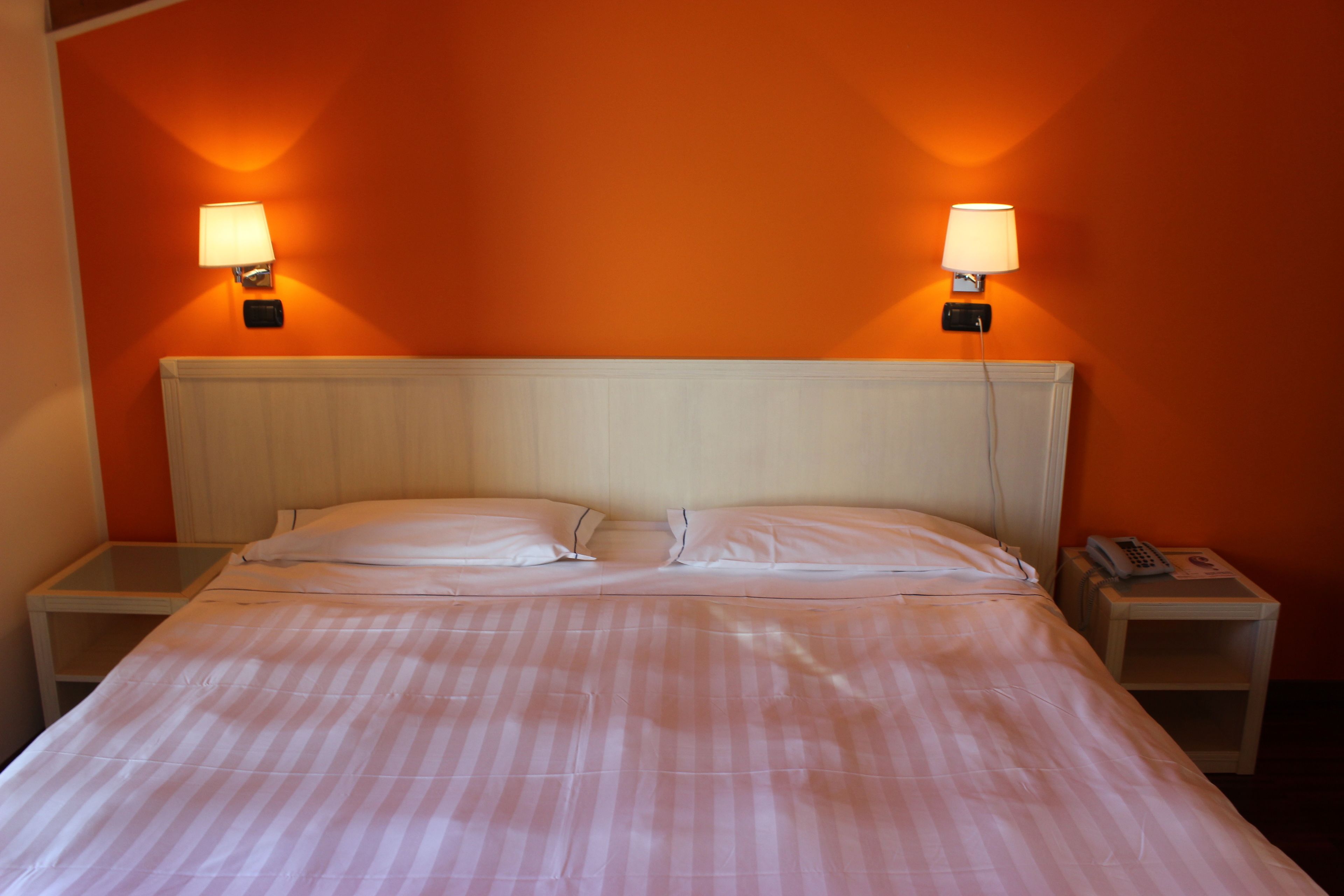 Quarto de hotel com uma cama, duas luminárias de cabeceira, uma mesinha de apoio com telefone e uma luminária na parede.
