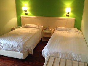 Deux lits simples avec du linge de lit blanc, une tête de lit en bois, des tables de chevet avec des lampes et un mur vert.