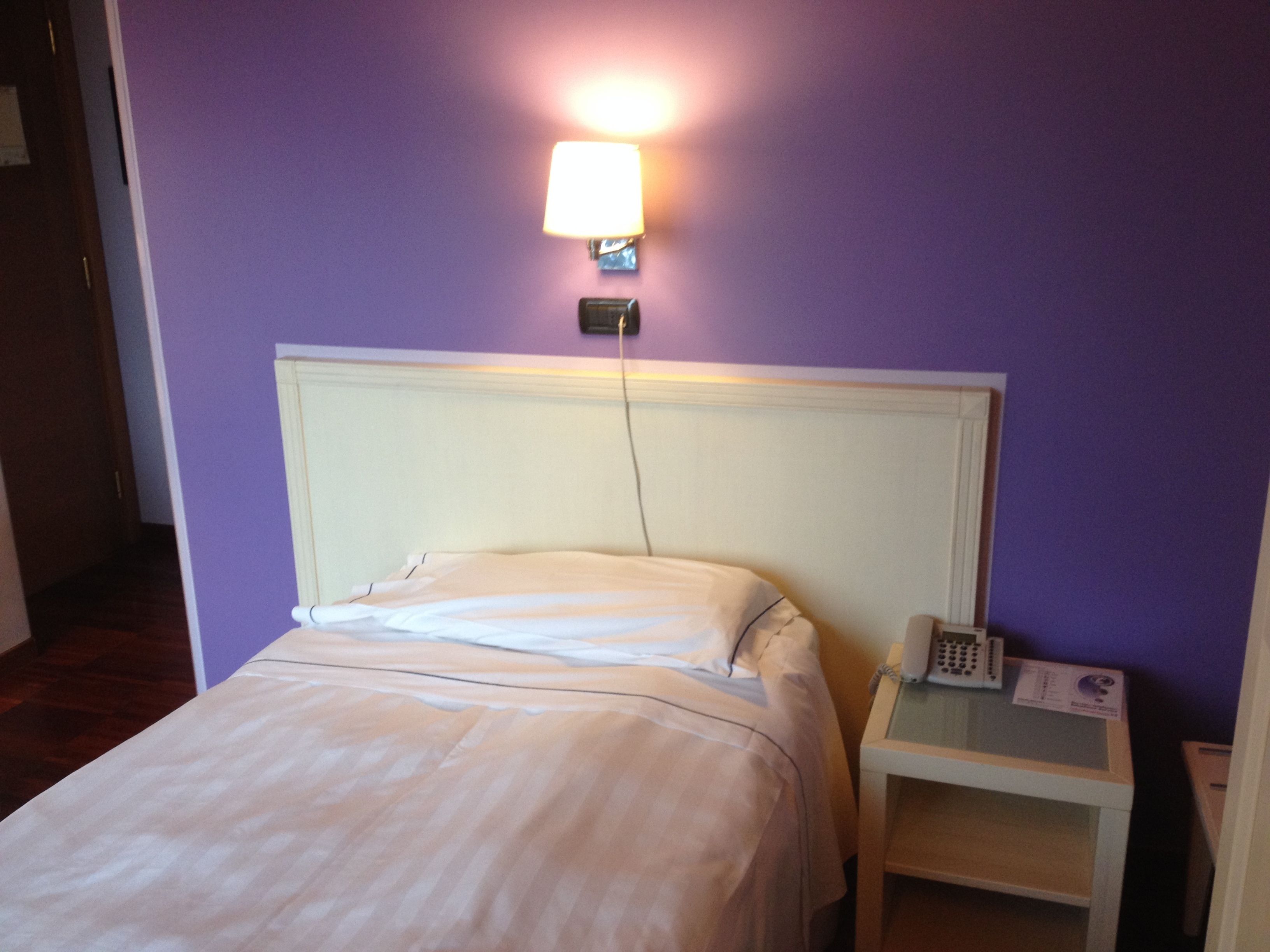 Habitación de hotel con una cama, una mesita de noche con un teléfono y una lámpara fijada a la pared.
