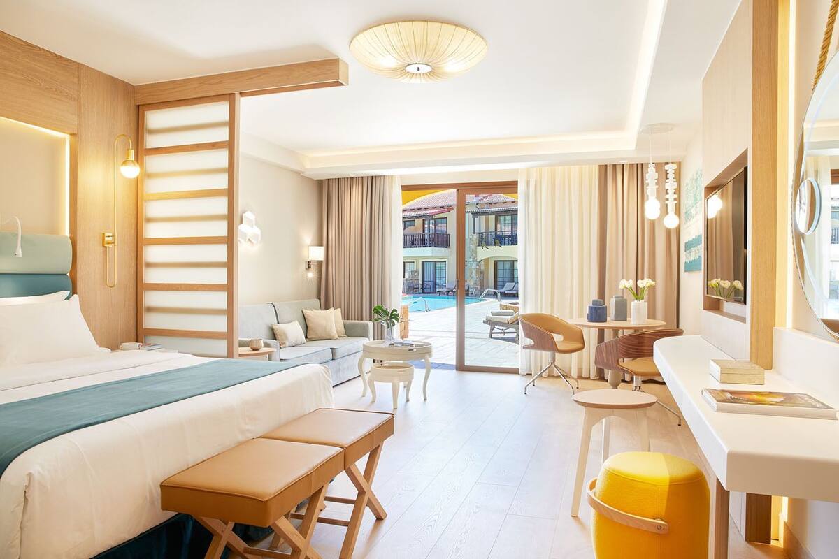 A hotel room with a bed, a desk, a chair, a small table, a bench, a mirror, and a view of the pool area.