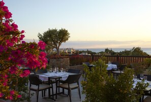 An outdoor dining area with tables set for two, surrounded by greenery and a view of the sea. - Anthemus Sea Beach Hotel & Spa (Sithonia)