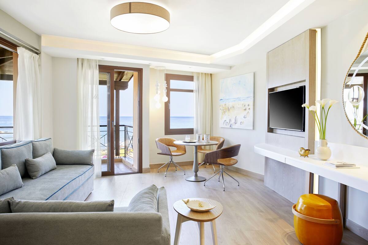 A modern hotel room with a sofa, dining table, chairs, a TV, and a view of the sea.