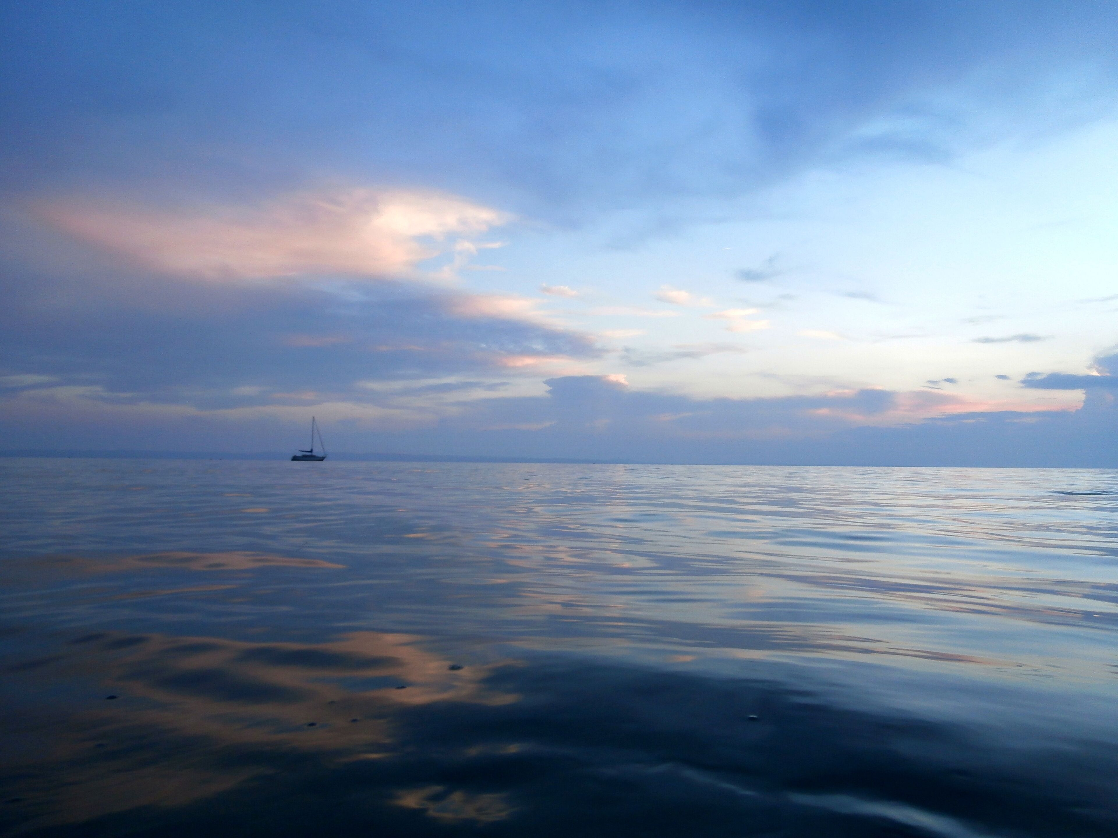 A calm sea with a sailboat in the distance under a cloudy sky.