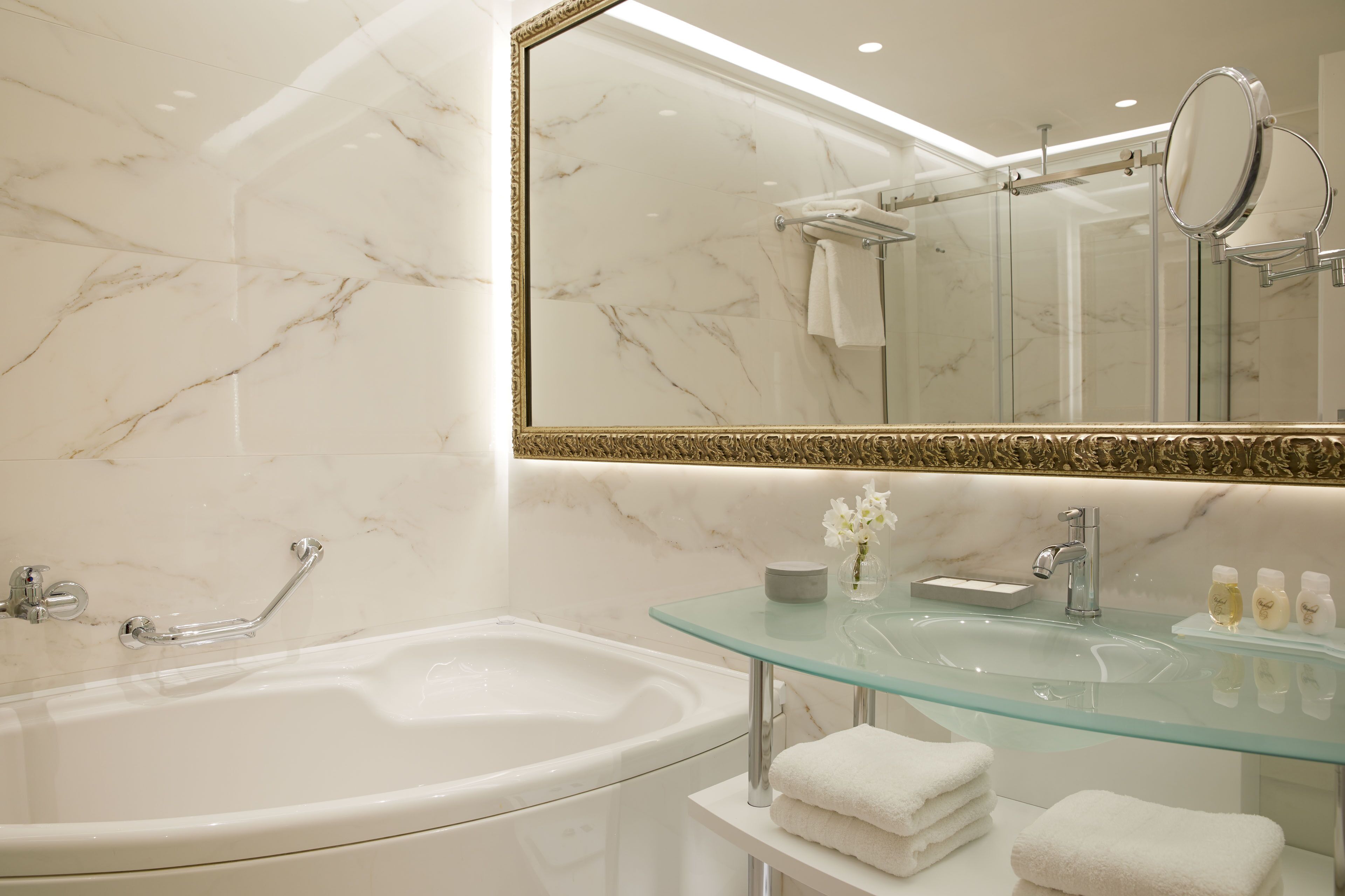 A modern bathroom with a marble wall, a glass sink, a bathtub, and a large mirror.