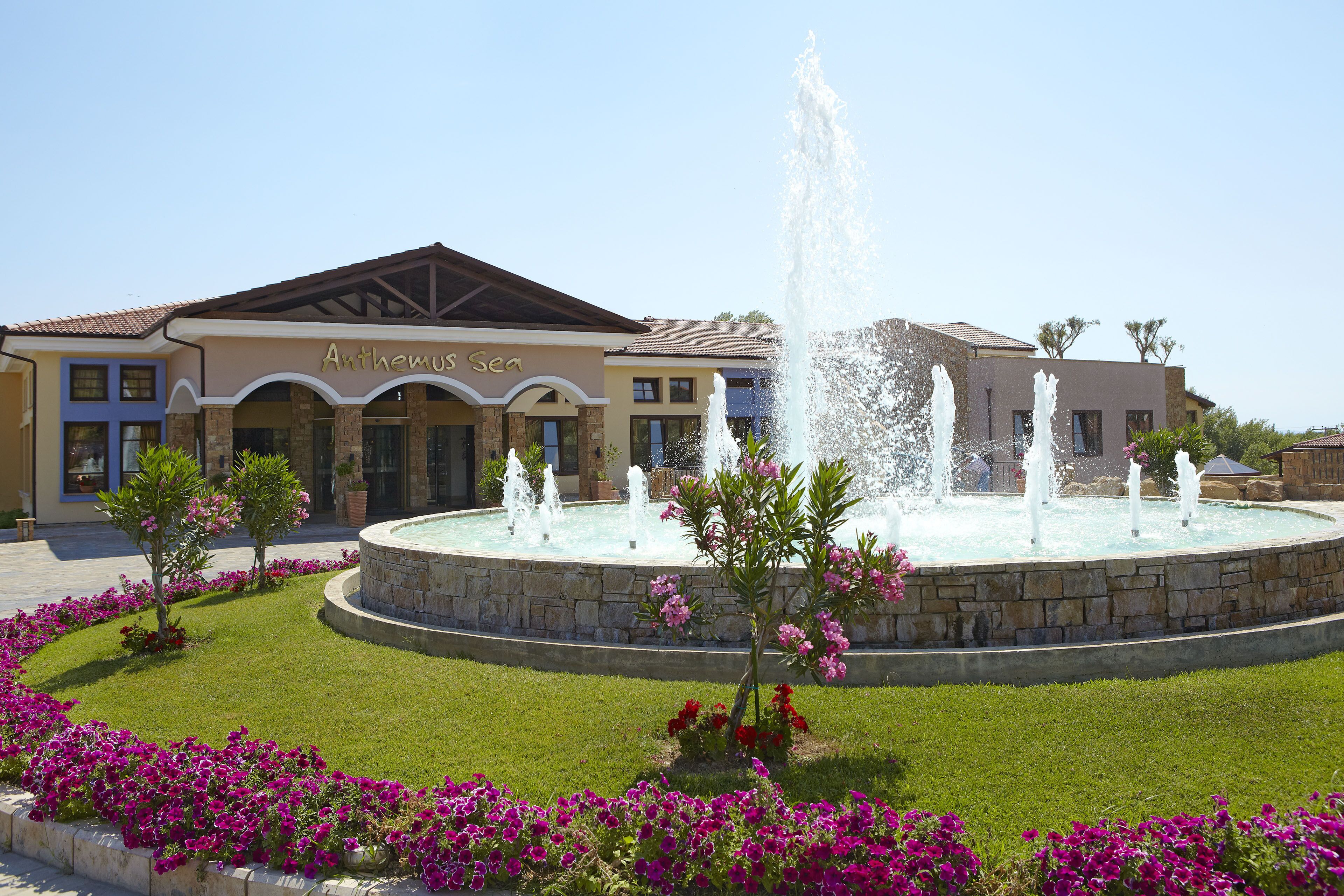 A hotel with a fountain in the center, surrounded by a well-maintained lawn and vibrant flowers.