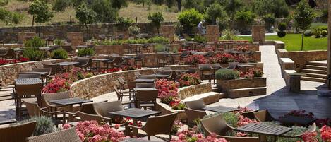 Un café al aire libre con paredes de piedra, mobiliario de mimbre y plantas en macetas.