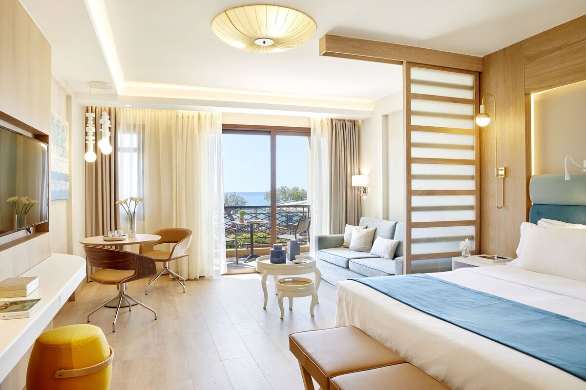 A hotel room with a bed, a sofa, a desk with chairs, a small table, and a balcony with a view.