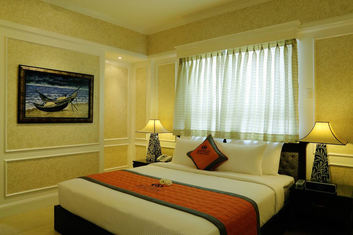 A hotel room with a large bed, two bedside tables, a nightstand, a window with curtains, a painting on the wall, and a lamp on each bedside table.