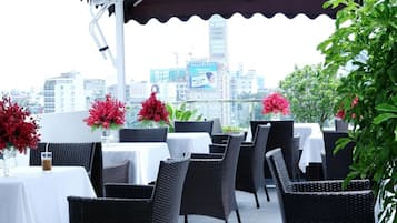 An outdoor dining area with wicker chairs and tables, red flowers, and a cityscape view in the background.