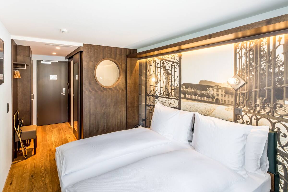 Habitación de hotel con una cama, un espejo, una puerta y vista a un paisaje urbano a través de un biombo metálico decorativo.