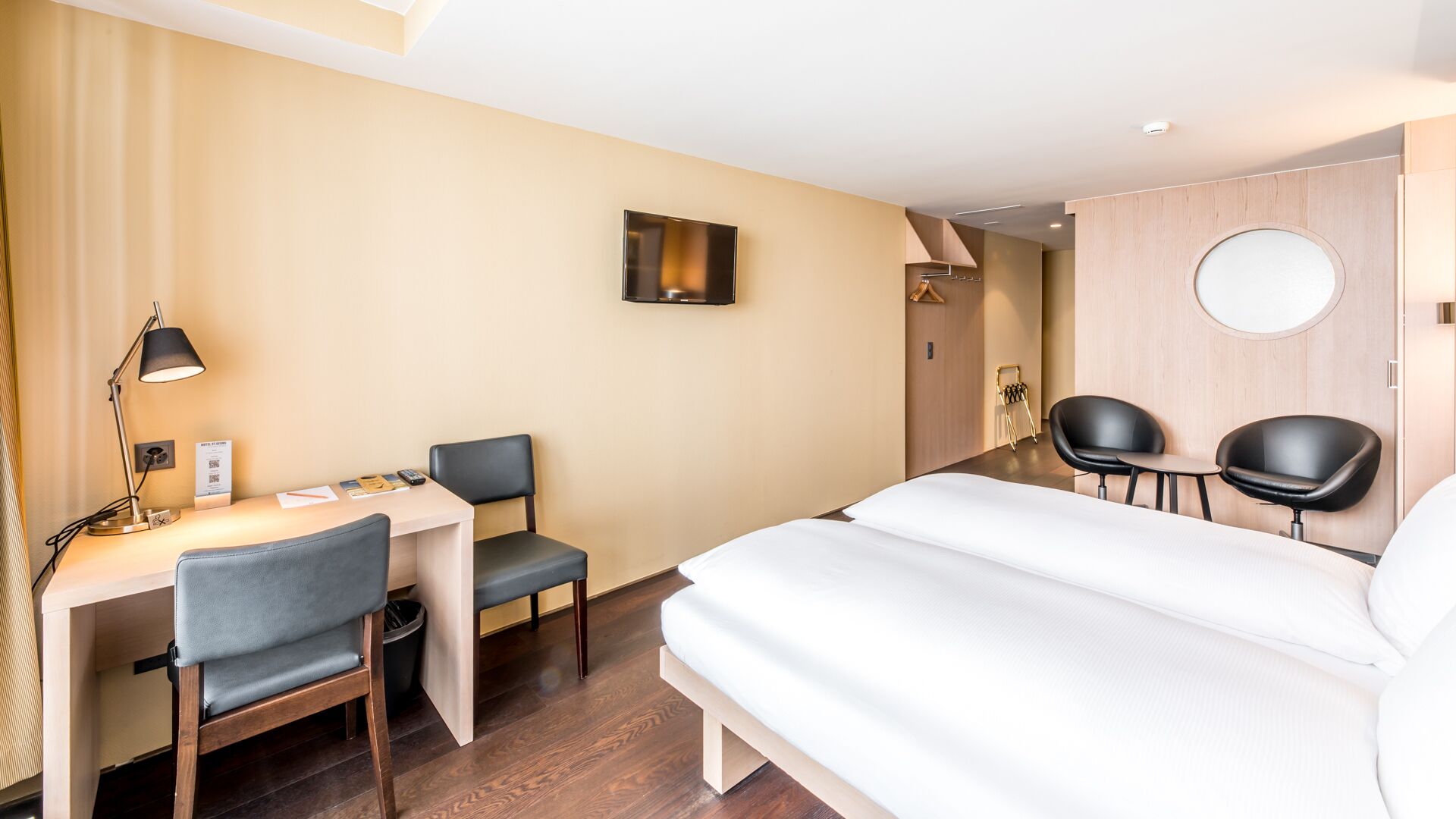 Une chambre d’hôtel équipée d’un lit, d’un bureau, de deux chaises, d’une lampe et d’un téléviseur fixé au mur.