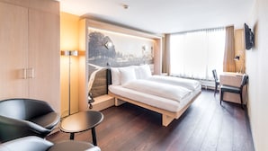 A modern hotel room with a bed, a desk, a chair, and a view of a cityscape through a large window. - Boutique Hotel St. Georg (Einsiedeln)