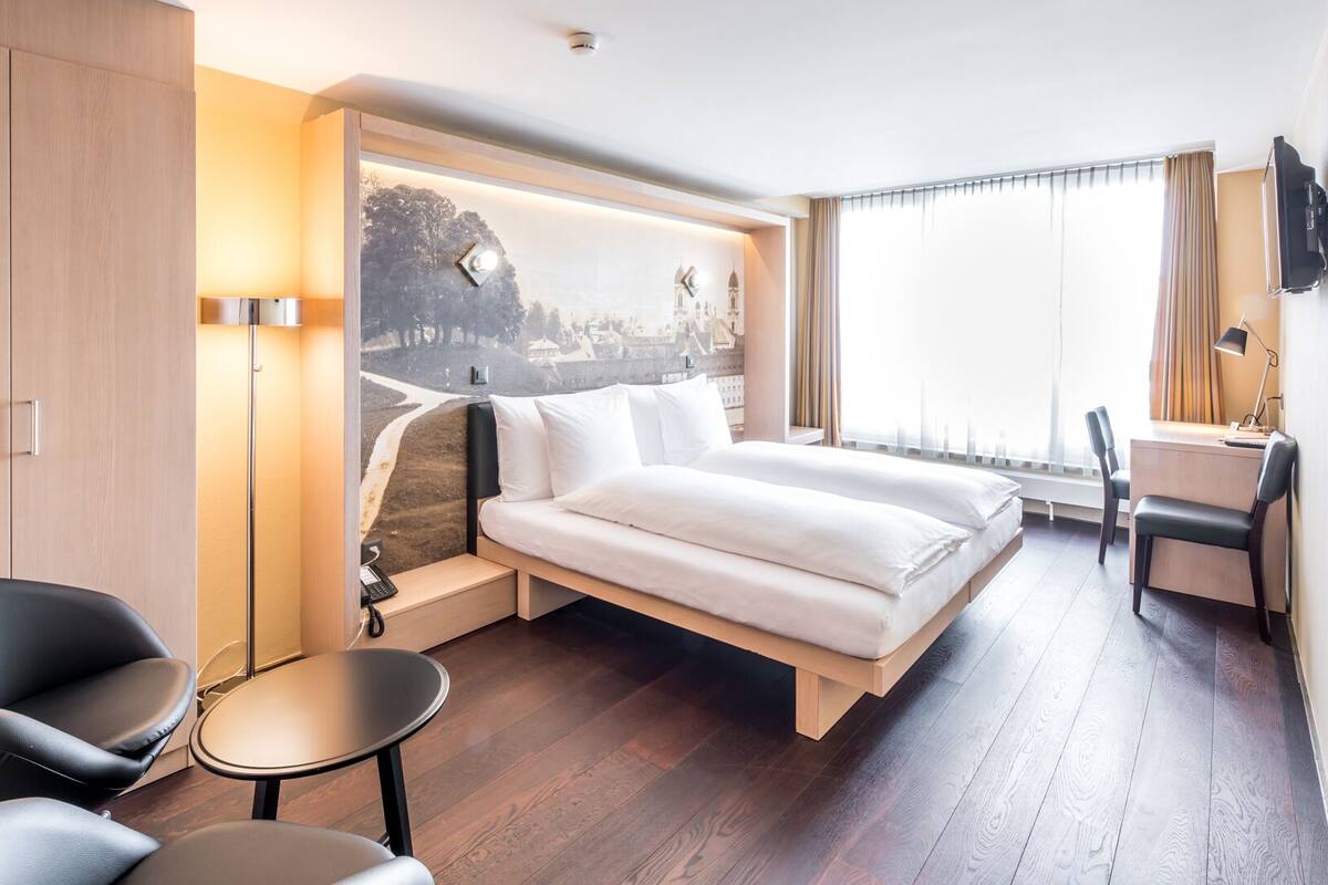 Habitación de hotel moderna con una cama, un escritorio, una silla y vistas a un paisaje urbano a través de un ventanal grande.