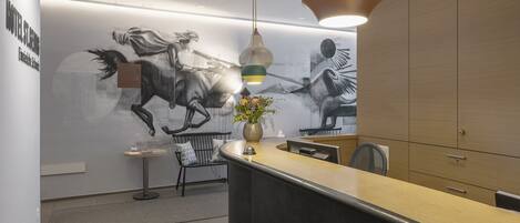A modern hotel lobby with a reception desk, a bench, a vase with flowers, and a wall mural of a horse race.