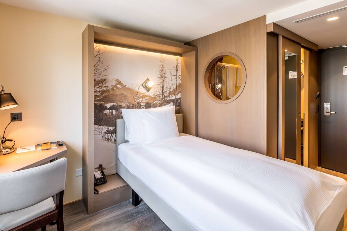 Habitación de hotel con cama, escritorio, silla, lámpara de escritorio, espejo y puerta.