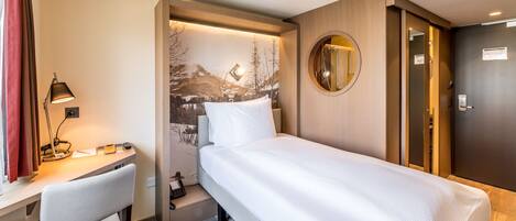 Habitación de hotel con cama, escritorio, silla, lámpara de escritorio, espejo y puerta.