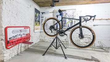 Una bicicleta en un soporte dentro de una habitación con pared de ladrillo blanco, un portaherramientas rojo y carteles.