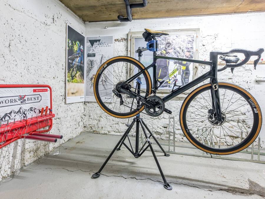 Una bicicleta en un soporte dentro de una habitación con pared de ladrillo blanco, un portaherramientas rojo y carteles.