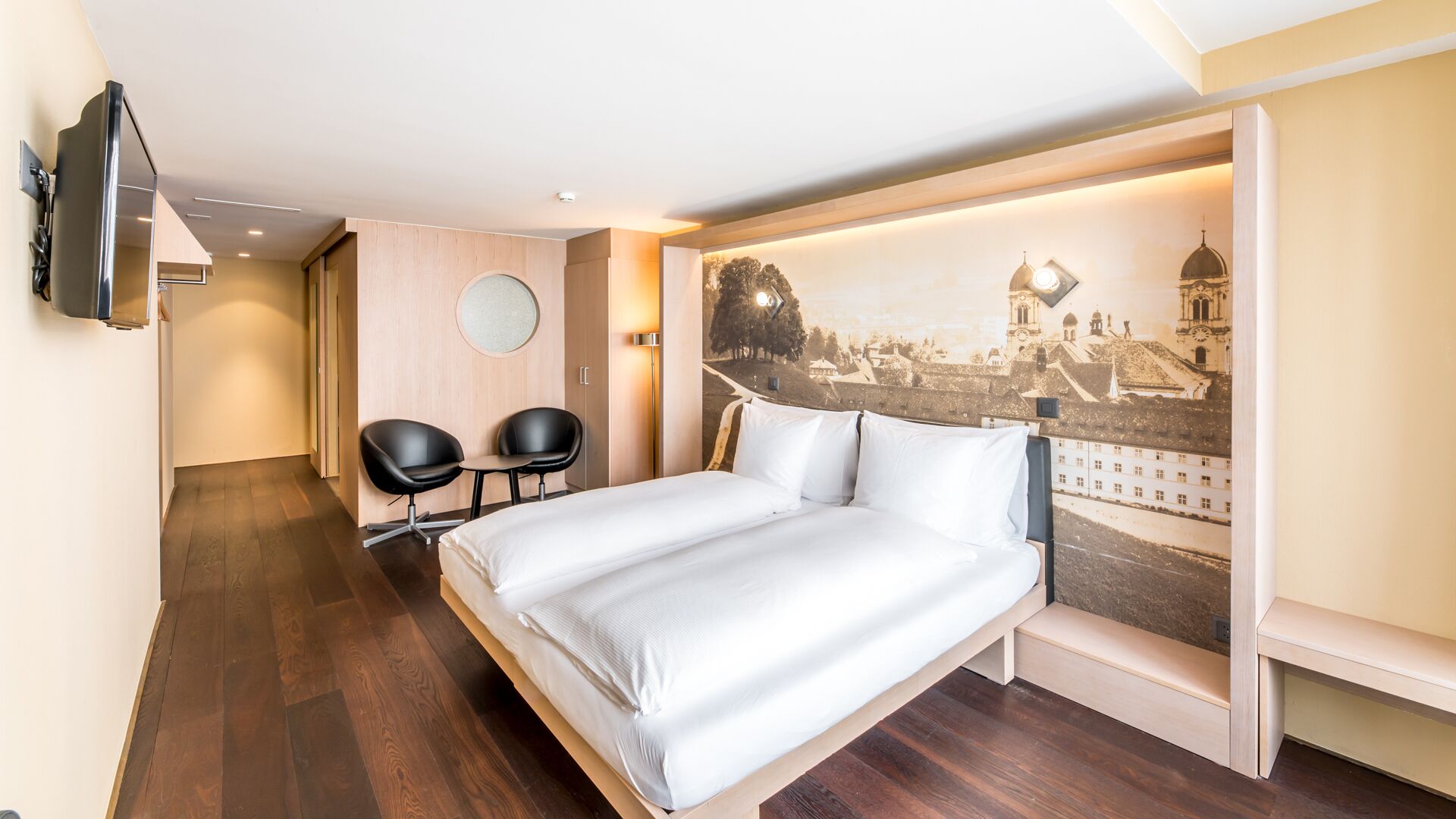 A hotel room with a bed, two black chairs, a TV, and a cityscape mural on the wall.