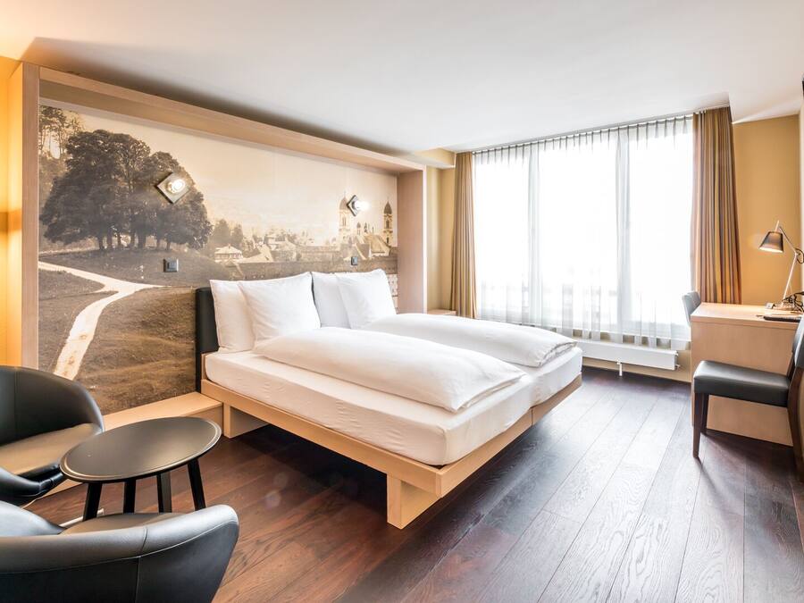 A hotel room with a bed, two chairs, a desk, a TV, and a large window with curtains.