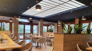 A modern restaurant with high ceilings, large windows, and a variety of seating arrangements. - Hotel le Caballin (Vogelgrun)