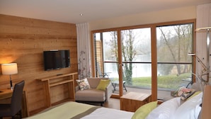A modern hotel room with a large bed, a TV mounted on a wooden wall, a desk with a chair, and a balcony with a view of a lake. - Hotel le Caballin (Vogelgrun)