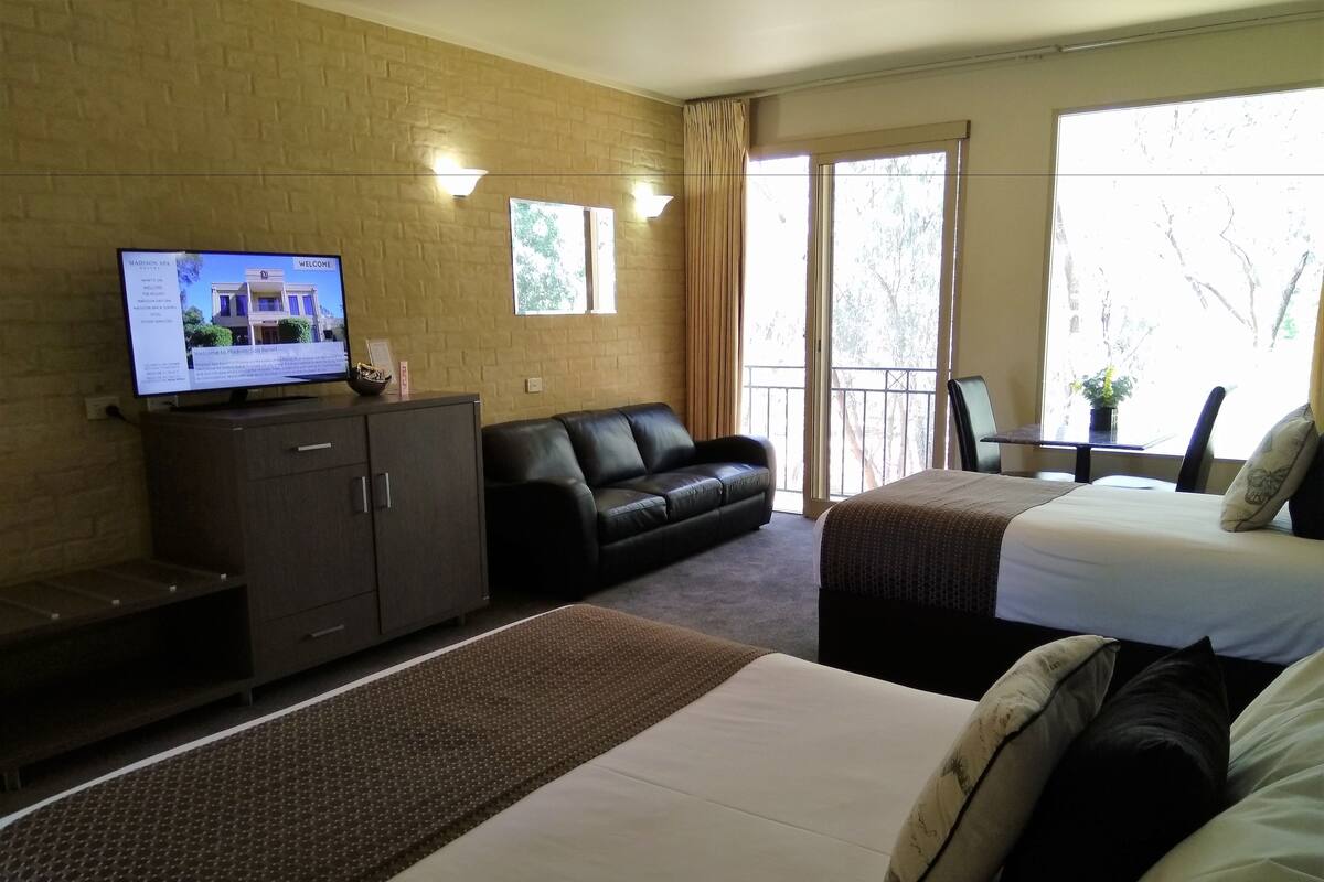 A hotel room with a bed, a sofa, a TV on a cabinet, and a balcony with a view of trees.