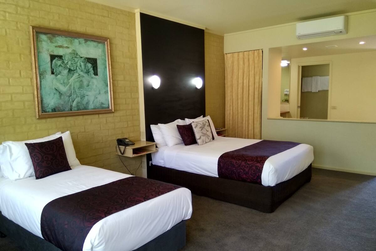 A hotel room with two beds, a painting on the wall, a bedside table with a phone, and an air conditioning unit.
