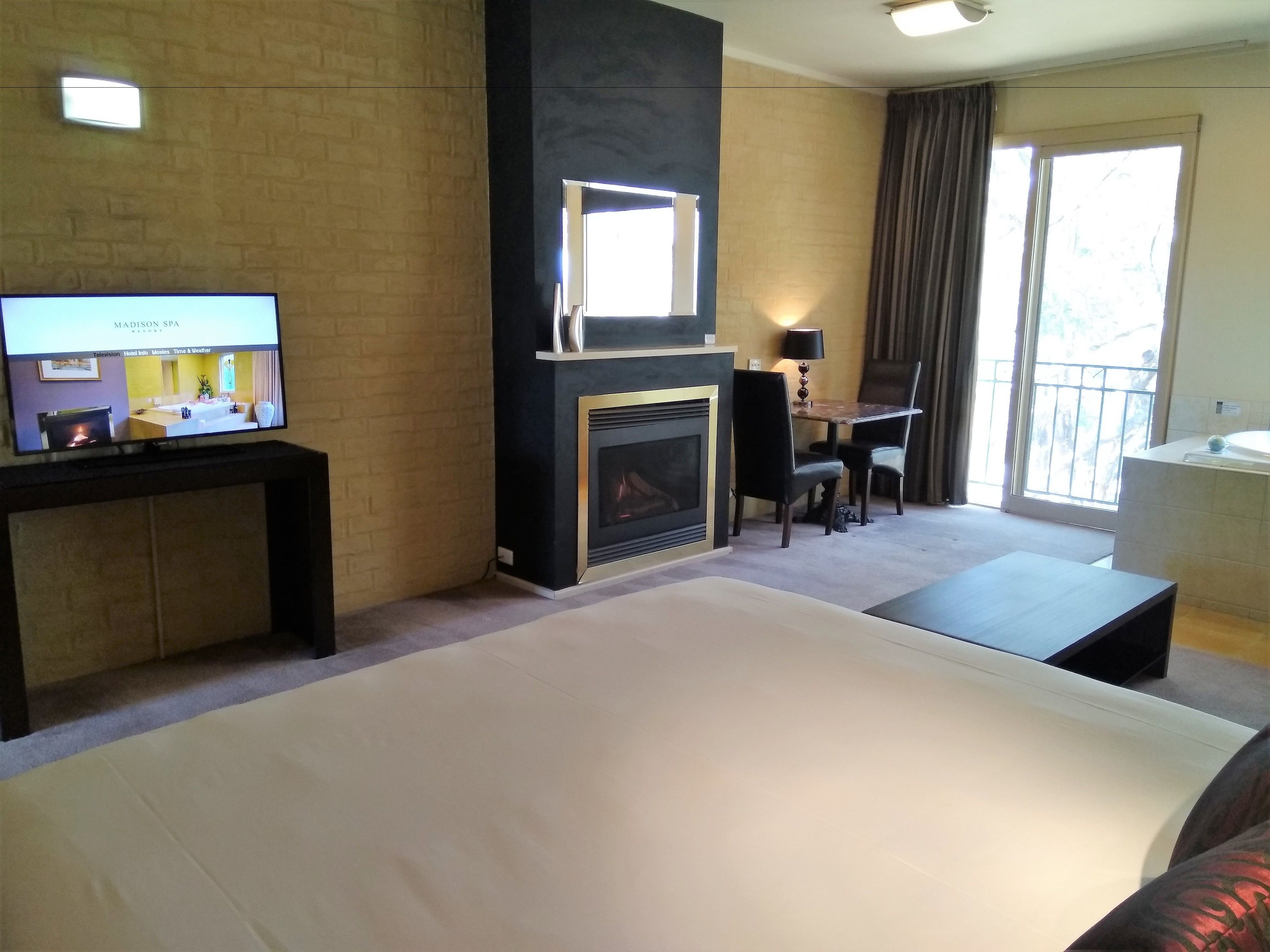 Habitación de hotel con cama, televisión mostrando un plano de la habitación, chimenea, zona de comedor con mesa y sillas, y ventana grande.