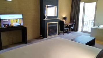Habitación de hotel con cama, televisión mostrando un plano de la habitación, chimenea, zona de comedor con mesa y sillas, y ventana grande.