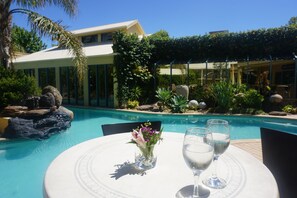 A poolside table with two glasses of water and a vase of flowers, surrounded by lush greenery and a clear blue pool. - Madison Spa Motel - Adults Only (Moama)