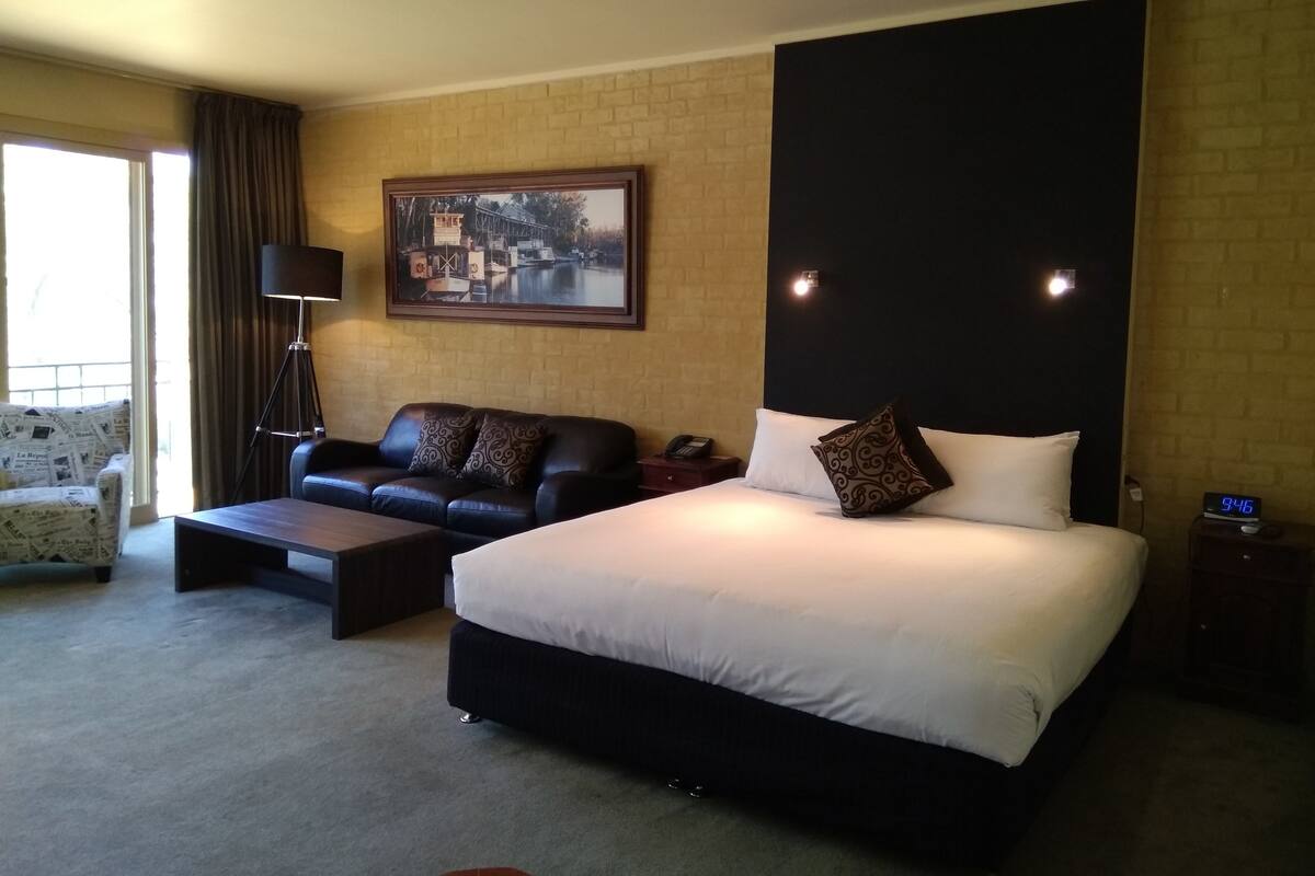A hotel room with a large bed, a sofa, a desk, a chair, a painting on the wall, and a clock on the bedside table.