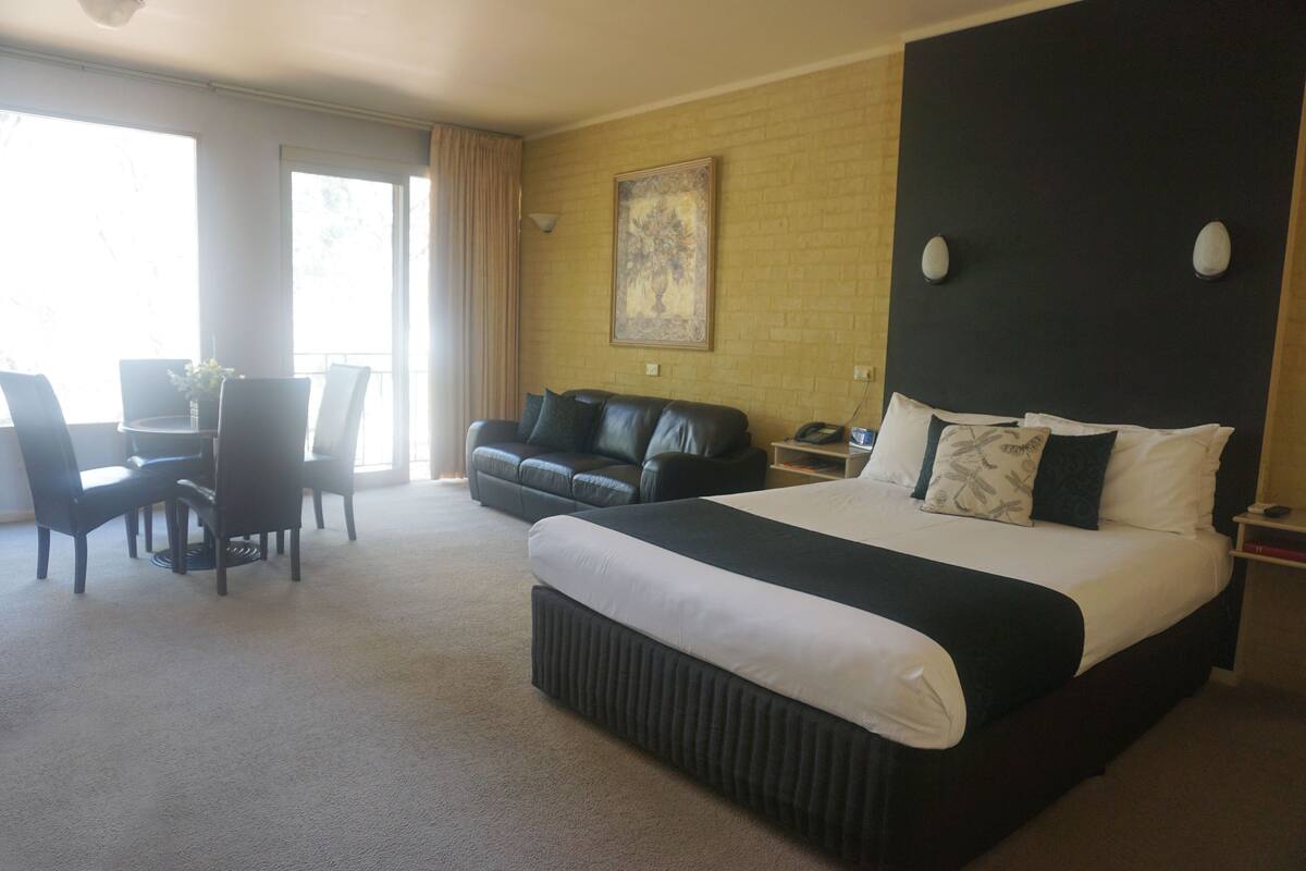 A hotel room with a large bed, a black leather sofa, a dining table with chairs, and a large window with curtains.