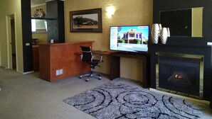 Un área de recepción con un escritorio de madera, una silla de oficina negra, un televisor mostrando un mensaje de bienvenida, una chimenea y una alfombra estampada.