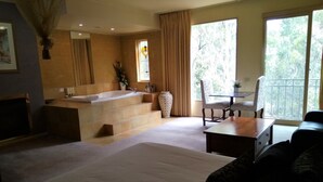 Una habitación amplia con una cama grande, un área de descanso con una mesa y sillas, una bañera y un ventanal con vistas a árboles.