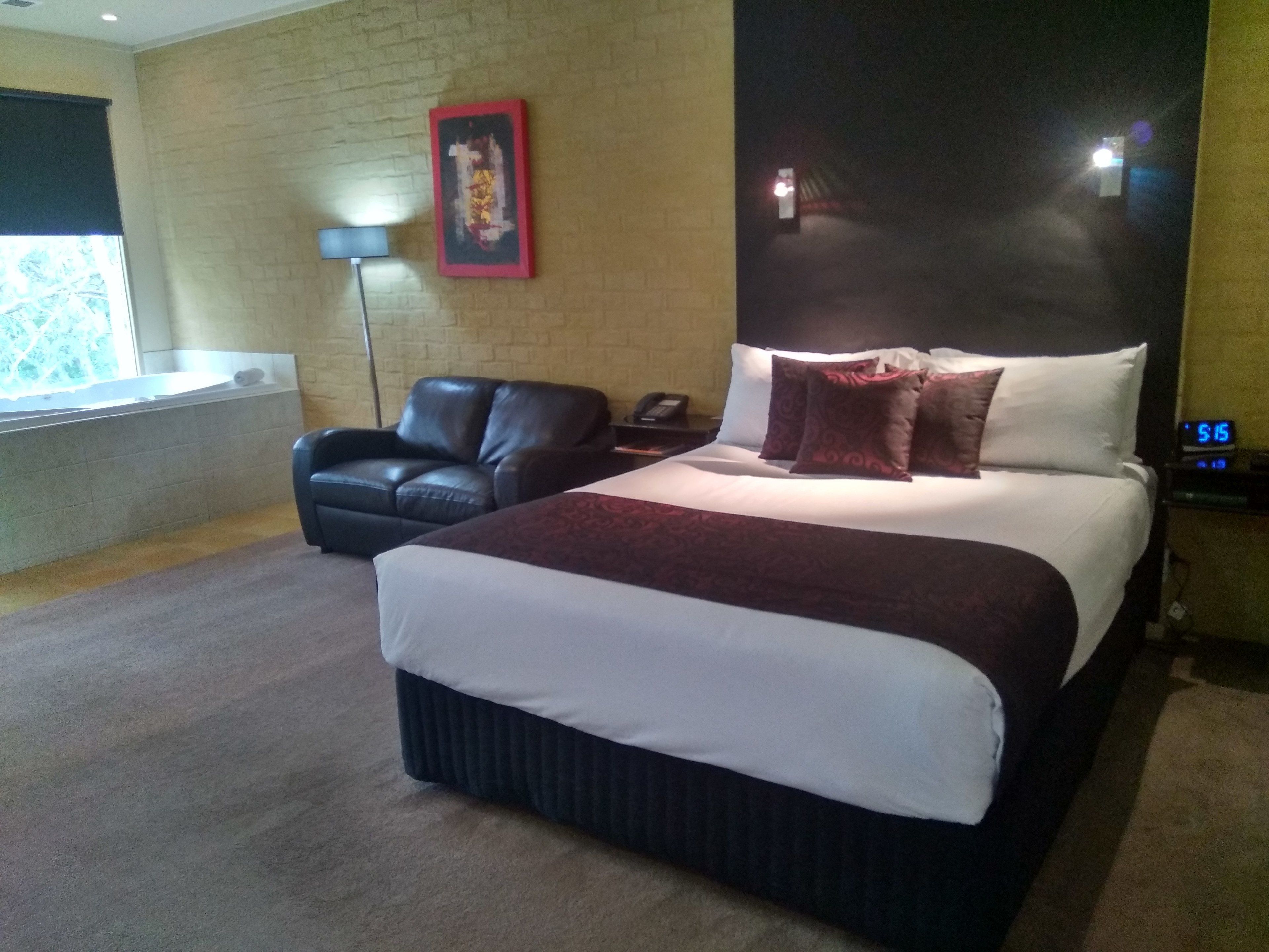 A hotel room with a large bed, a leather chair, a bedside table with a lamp, and a wall clock.