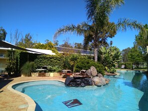 A swimming pool with a rock feature, surrounded by palm trees and a covered patio area. - Madison Spa Motel - Adults Only (Moama)