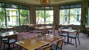 Un restaurante con varias mesas y sillas, amplias ventanas con vistas a zonas verdes y lámparas colgantes.