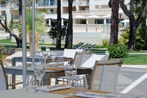 An outdoor dining area with tables and chairs, a palm tree, and a building in the background. - Hotel Don Cesar (Porto-Vecchio)