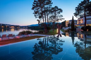 A serene evening poolside area with lounge chairs, a clear reflection in the water, and a backdrop of trees and distant buildings. - Hotel Don Cesar (Porto-Vecchio)