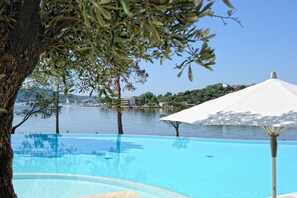 An infinity pool with a clear view of the water and distant buildings, surrounded by trees and a white umbrella. - Hotel Don Cesar (Porto-Vecchio)