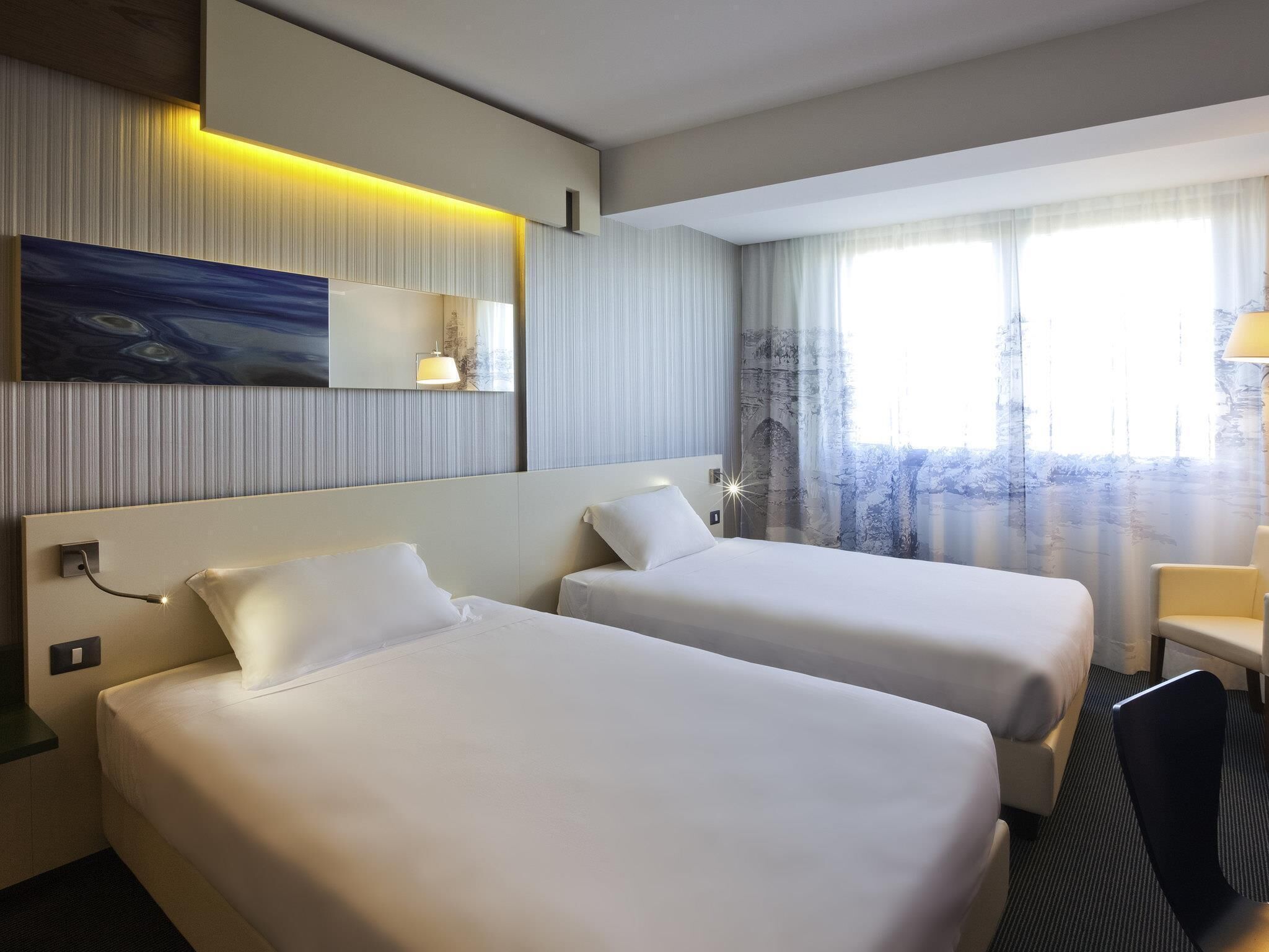 A hotel room with two beds, a painting on the wall, a window with curtains, and a lamp.