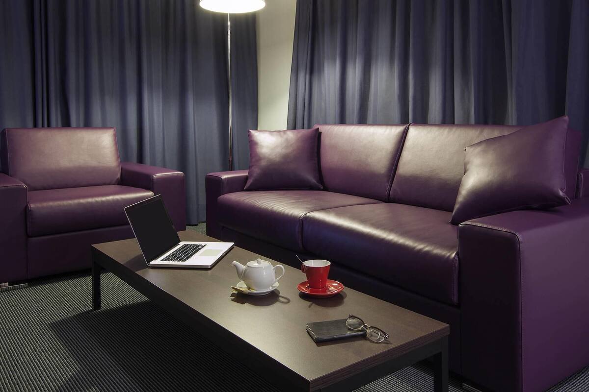 A modern hotel room with a sofa, a coffee table with a laptop, a teapot, a red cup, and a pair of glasses.