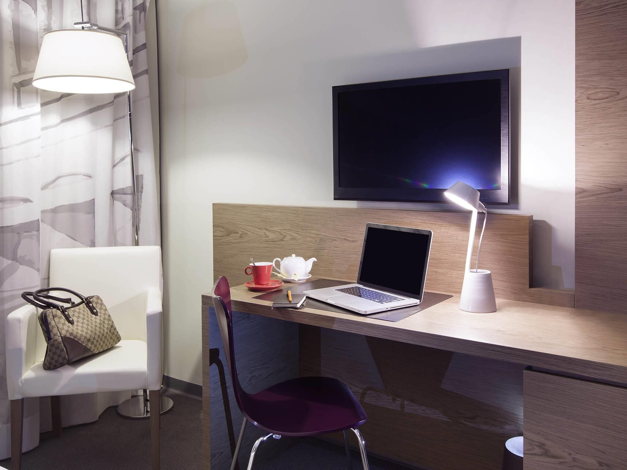 Une chambre d’hôtel équipée d’un bureau, d’un ordinateur portable, d’une télévision, d’une chaise, d’un sac à main et d’une lampe.