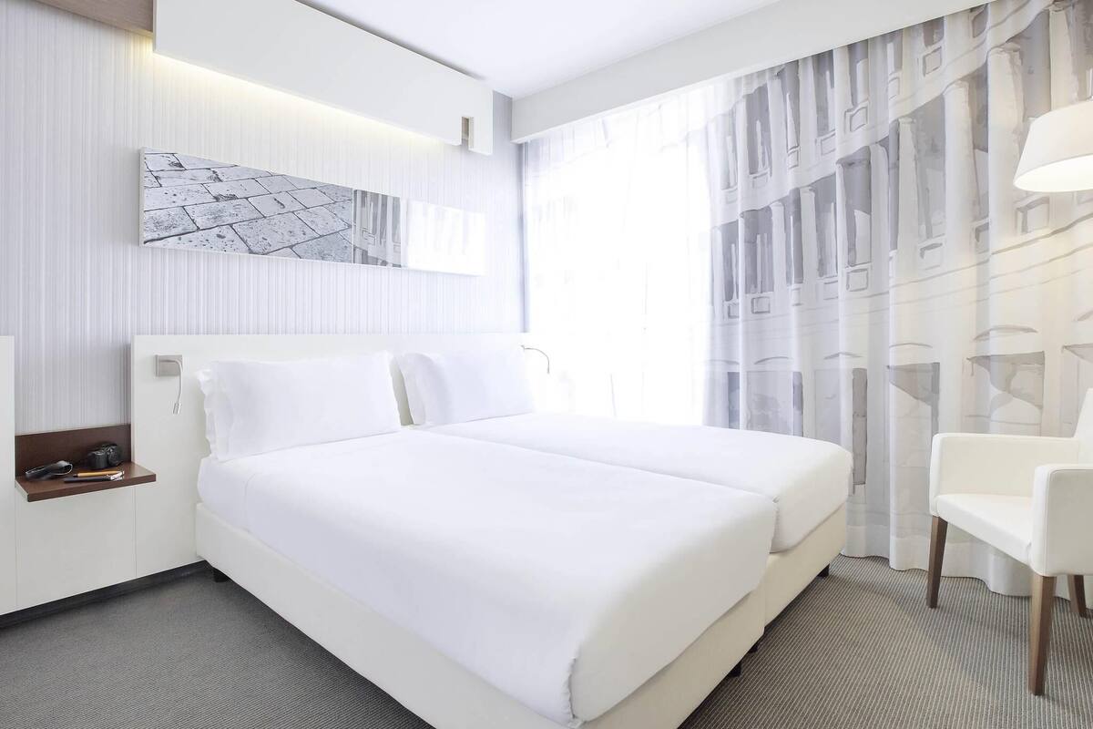 A hotel room with a large bed, a bedside table with a lamp, a chair, and a window with sheer curtains.