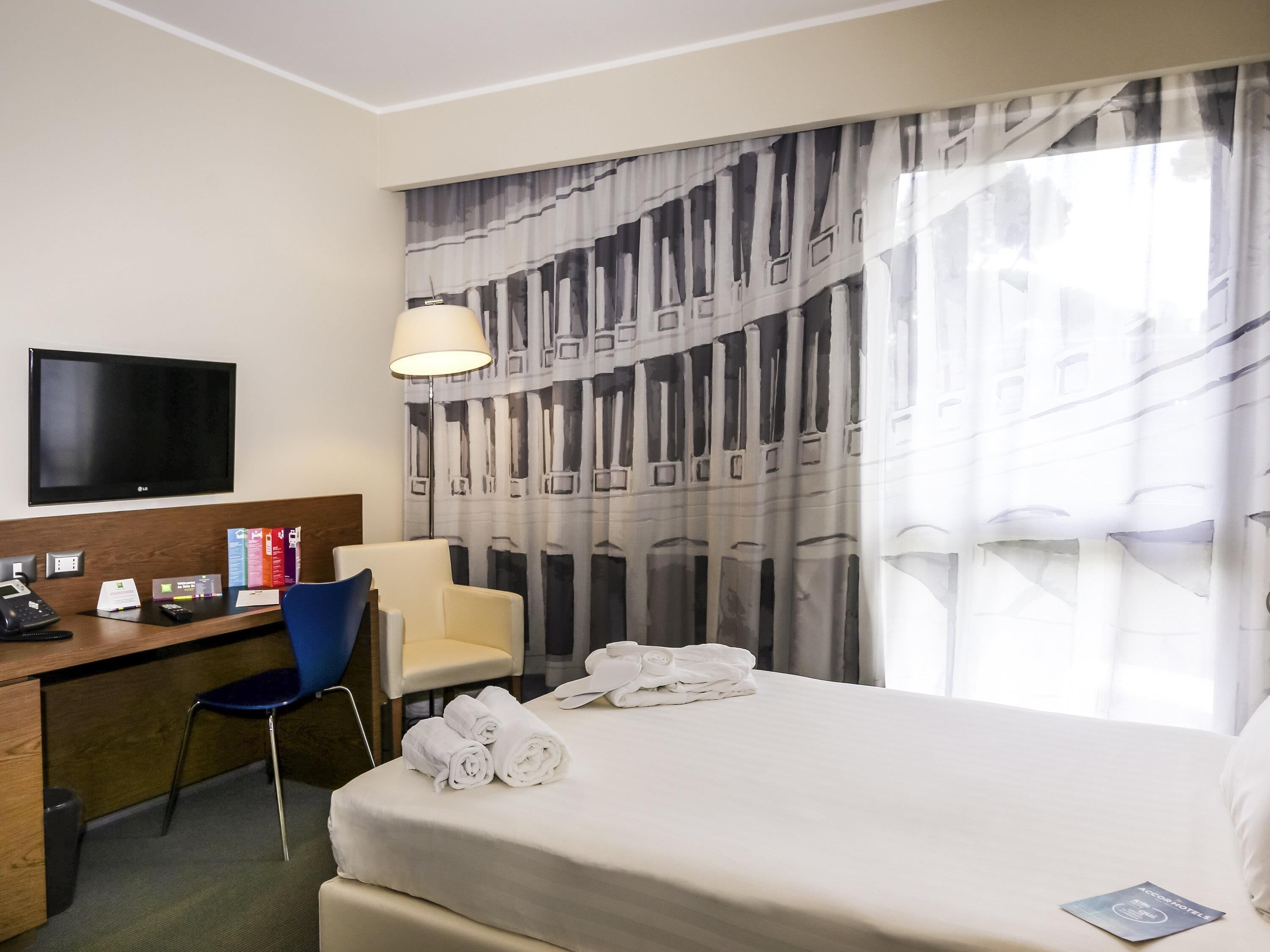 Quarto de hotel com cama, escrivaninha, televisão de tela plana, cadeira e janela com cortinas.